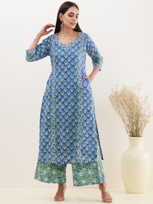 A woman is wearing a traditional blue and green patterned kurta and palazzo set. She stands with one hand near her face and the other in a pocket. The outfit features floral motifs and is complemented by tan sandals and green earrings. In the background, there is a light beige wall and a large ceramic vase with dried plants.