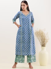 A woman is wearing a blue and green patterned kurta with matching palazzo pants, standing with hands in her pockets. She accessorizes with green earrings and sandals. In the background, there is a tall cream-colored vase with dried grass.