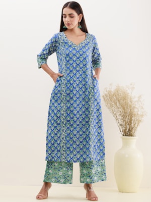 A woman is wearing a blue and green patterned kurta with matching palazzo pants, standing with hands in her pockets. She accessorizes with green earrings and sandals. In the background, there is a tall cream-colored vase with dried grass.