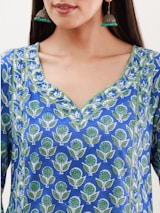A woman wearing a blue kurta featuring a floral pattern with green and white accents. She has long hair and is wearing large, ornate earrings with a gold finish and turquoise beads.