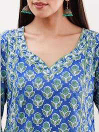 A woman wearing a blue kurta featuring a floral pattern with green and white accents. She has long hair and is wearing large, ornate earrings with a gold finish and turquoise beads.