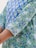 A close-up view of a garment featuring a floral and leafy print in shades of blue and green. The intricate design includes small flowers and abstract foliage, creating a vibrant and dynamic pattern on the fabric. The edge of the sleeve is detailed with a decorative trim.
