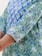 A close-up view of a garment featuring a floral and leafy print in shades of blue and green. The intricate design includes small flowers and abstract foliage, creating a vibrant and dynamic pattern on the fabric. The edge of the sleeve is detailed with a decorative trim.