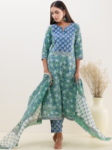 A woman is wearing a floral patterned blue and green kurta set, consisting of a long tunic and matching bottoms. The outfit is complemented by a flowing dupatta. Her long dark hair is straight and she is wearing earrings. A beige vase with dried plants is placed in the background.