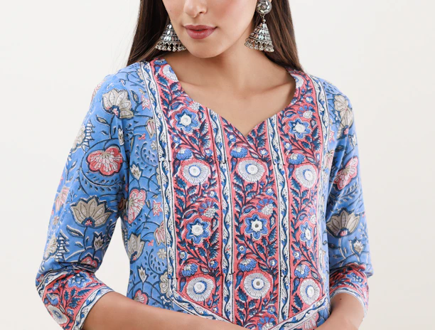 A person is wearing a blue floral-patterned kurta with intricate designs in shades of blue, red, and white. The attire has a v-shaped neckline and three-quarter-length sleeves. They are accessorized with large, ornate, silver jhumka earrings. The hair is styled straight and parted in the middle.