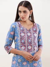 A person is wearing a blue floral-patterned kurta with intricate designs in shades of blue, red, and white. The attire has a v-shaped neckline and three-quarter-length sleeves. They are accessorized with large, ornate, silver jhumka earrings. The hair is styled straight and parted in the middle.