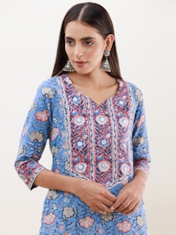 A person is wearing a blue floral-patterned kurta with intricate designs in shades of blue, red, and white. The attire has a v-shaped neckline and three-quarter-length sleeves. They are accessorized with large, ornate, silver jhumka earrings. The hair is styled straight and parted in the middle.