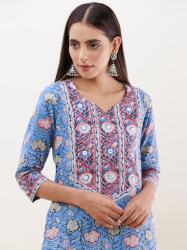 A person is wearing a blue floral-patterned kurta with intricate designs in shades of blue, red, and white. The attire has a v-shaped neckline and three-quarter-length sleeves. They are accessorized with large, ornate, silver jhumka earrings. The hair is styled straight and parted in the middle.
