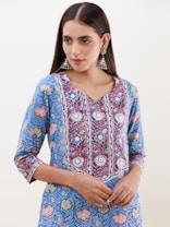 A person is wearing a blue floral-patterned kurta with intricate designs in shades of blue, red, and white. The attire has a v-shaped neckline and three-quarter-length sleeves. They are accessorized with large, ornate, silver jhumka earrings. The hair is styled straight and parted in the middle.