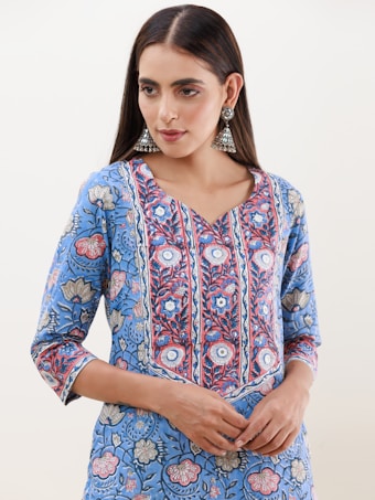 A person is wearing a blue floral-patterned kurta with intricate designs in shades of blue, red, and white. The attire has a v-shaped neckline and three-quarter-length sleeves. They are accessorized with large, ornate, silver jhumka earrings. The hair is styled straight and parted in the middle.