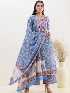 A woman wearing a traditional floral-patterned blue and white dress with intricate designs stands with her hands clasped in front of her. She is accessorized with large silver earrings. To her left, there is a large white vase containing tall, dried plants.