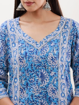 A close-up view of a person wearing a blue floral patterned garment with intricate designs. The person is accessorized with large, metallic jhumka earrings and has long hair. The focus is on the upper body, showing the vivid details of the fabric pattern.