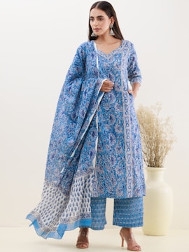 A person is wearing a blue traditional outfit with intricate floral patterns. The attire includes a long tunic, wide-legged pants, and a matching dupatta. The person is standing against a plain beige background with a large white vase containing dried grasses positioned on the right.