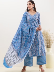 A woman is wearing a traditional blue patterned outfit, consisting of a long tunic and matching wide-legged pants. She is accessorized with large silver earrings and stands in front of a neutral background with a decorative vase containing dried flowers.