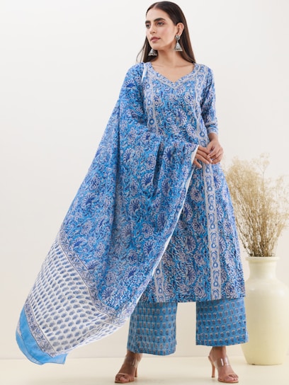 A woman is wearing a traditional blue patterned outfit, consisting of a long tunic and matching wide-legged pants. She is accessorized with large silver earrings and stands in front of a neutral background with a decorative vase containing dried flowers.