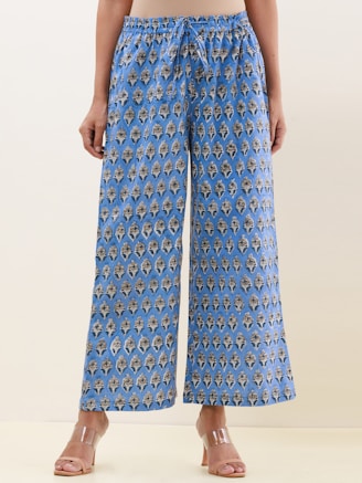 A pair of wide-legged pants featuring an intricate, symmetrical pattern design in blue with darker and lighter contrasts. The fabric gives a casual yet sophisticated appearance, and the pants are paired with clear, modern-looking heeled sandals. The background is a neutral shade, highlighting the pants.