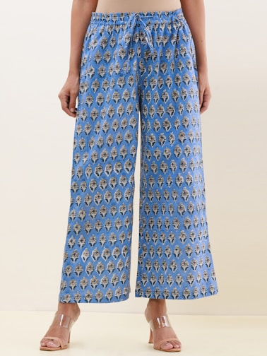 A pair of wide-legged pants featuring an intricate, symmetrical pattern design in blue with darker and lighter contrasts. The fabric gives a casual yet sophisticated appearance, and the pants are paired with clear, modern-looking heeled sandals. The background is a neutral shade, highlighting the pants.
