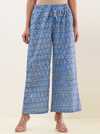 A pair of wide-legged pants featuring an intricate, symmetrical pattern design in blue with darker and lighter contrasts. The fabric gives a casual yet sophisticated appearance, and the pants are paired with clear, modern-looking heeled sandals. The background is a neutral shade, highlighting the pants.