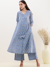 A woman is wearing a blue patterned kurta with matching palazzo pants, accompanied by large silver earrings. Her hair is straight and she stands against a neutral background, which includes a decorative vase with dried flowers.