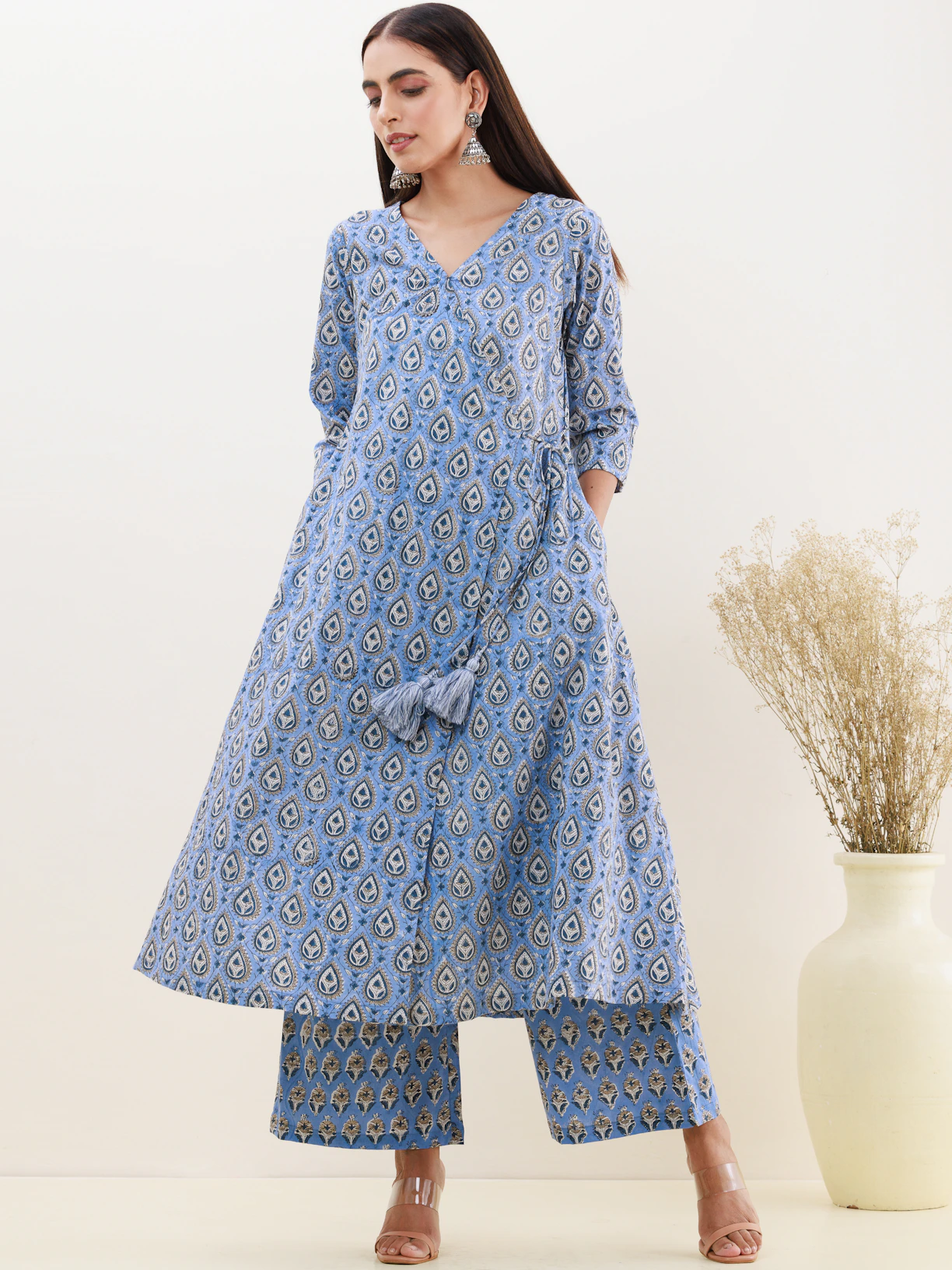 A woman is wearing a blue patterned kurta with matching palazzo pants, accompanied by large silver earrings. Her hair is straight and she stands against a neutral background, which includes a decorative vase with dried flowers.