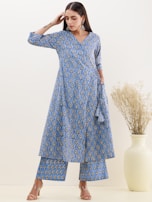 A person is wearing a matching blue patterned kurta and pants set, with intricate designs and a relaxed fit. The outfit includes a long kurta with a V-neck and three-quarter sleeves. The person accessorizes with large silver earrings and open-toe heels. A light background with a large decorative vase containing dried plants adds to the composition.