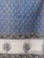 An intricate textile design featuring a repeating pattern of teardrop-shaped motifs on a blue background. The motifs contain detailed floral and leaf patterns, bordered by a series of elaborate floral designs. The bottom section features larger floral motifs on a white background, framed by a floral border.