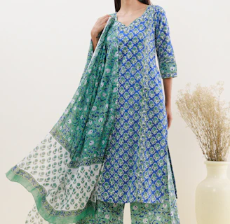 A person is wearing a blue and green floral patterned traditional outfit. The attire includes a long kurta, matching pants, and a dupatta with varying patterns. The background is a neutral off-white color, and there is a large ceramic vase with dried grass on the right.