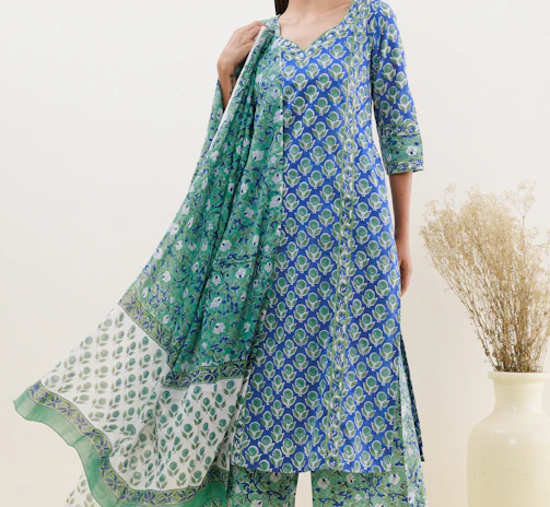 A person is wearing a blue and green floral patterned traditional outfit. The attire includes a long kurta, matching pants, and a dupatta with varying patterns. The background is a neutral off-white color, and there is a large ceramic vase with dried grass on the right.