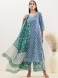 A person is wearing a blue and green floral patterned traditional outfit. The attire includes a long kurta, matching pants, and a dupatta with varying patterns. The background is a neutral off-white color, and there is a large ceramic vase with dried grass on the right.