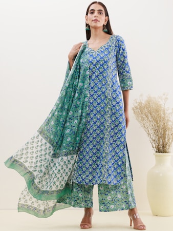A person is wearing a blue and green floral patterned traditional outfit. The attire includes a long kurta, matching pants, and a dupatta with varying patterns. The background is a neutral off-white color, and there is a large ceramic vase with dried grass on the right.