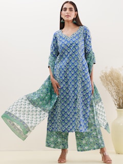 A person is wearing a blue and green floral printed outfit consisting of a long kurta and palazzo pants. The outfit has a traditional look with intricate patterns. The person is posing confidently in front of a light beige background and is accessorized with green earrings.