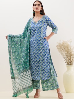A woman wearing a blue and green floral-patterned traditional outfit, including a long kurta, printed palazzo pants, and a matching dupatta. She's posing with one hand on her hip and the other holding the edge of the dupatta. The background is minimalist, featuring a light wall and a vase with dried grass.
