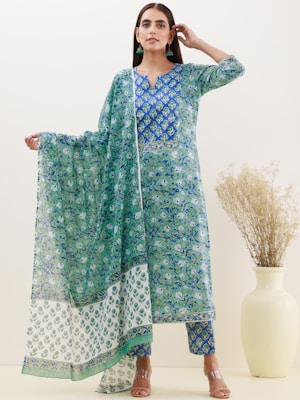 A woman is wearing a traditional outfit featuring an intricate blue and green floral pattern. The ensemble includes a long kurta, pants, and a matching dupatta. The woman has long dark hair and is accessorizing with earrings. A cream-colored vase with dried flowers is placed to her side.