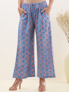A pair of wide-legged blue pants featuring a floral pattern with red and pink flowers. The pants have an elastic waistband and are worn with nude heels. Dry decorative plants are visible in a vase to the side.