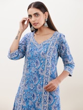 A person with long dark hair is wearing a blue, intricately patterned kurta. The kurta features floral designs and vertical borders, creating a traditional and elegant look. The individual has large silver earrings and a tattoo on their forearm. The expression is calm and poised, with a light background enhancing the focus on the person.