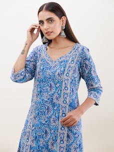 A person with long dark hair is wearing a blue, intricately patterned kurta. The kurta features floral designs and vertical borders, creating a traditional and elegant look. The individual has large silver earrings and a tattoo on their forearm. The expression is calm and poised, with a light background enhancing the focus on the person.