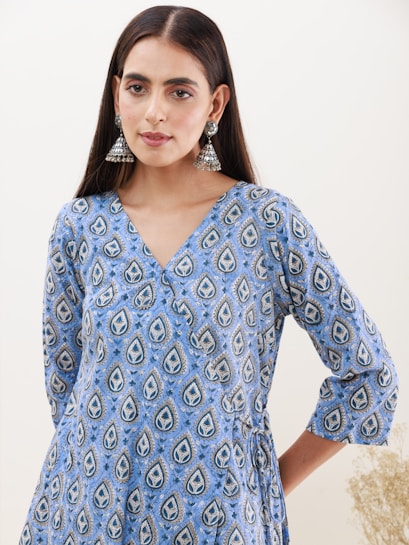 A woman with long dark hair is wearing a blue patterned dress adorned with a print consisting of teardrop designs. She has silver jhumka-style earrings and is posed in front of a plain background. The dress features a V-neck and three-quarter sleeves.