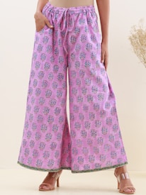 A person wearing wide-legged pants with a pink base and blue floral patterns. The pants have a drawstring waist and are paired with beige open-toed heels. The background features a light, neutral shade with some dried plant elements on the side.
