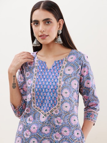 A person with long dark hair is wearing an intricately patterned tunic featuring floral designs in shades of blue, pink, and purple. Silver jhumka earrings with detailed craftsmanship complement the outfit. A small tattoo is visible on the person's forearm, and they have a subtle, neutral expression.