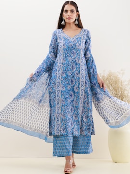 A woman is wearing a blue floral patterned ethnic outfit. She stands against a plain background while holding the edges of her dress, showcasing the intricate designs. She wears silver earrings and is barefoot, standing next to a white vase containing dried twigs.