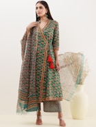 A woman stands modeling a traditional outfit featuring a long, green floral printed dress with intricate detailing and red tassels. She holds a sheer dupatta and wears matching pants. Her hair is long and straight, and she accessorizes with green earrings. A white vase with dry grass adds a decorative element to the background.