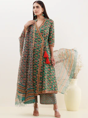 A woman stands modeling a traditional outfit featuring a long, green floral printed dress with intricate detailing and red tassels. She holds a sheer dupatta and wears matching pants. Her hair is long and straight, and she accessorizes with green earrings. A white vase with dry grass adds a decorative element to the background.