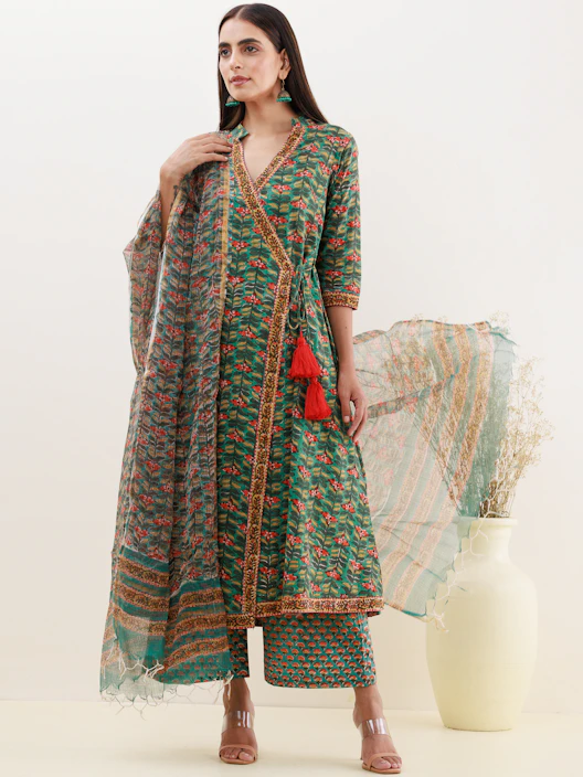 A woman stands modeling a traditional outfit featuring a long, green floral printed dress with intricate detailing and red tassels. She holds a sheer dupatta and wears matching pants. Her hair is long and straight, and she accessorizes with green earrings. A white vase with dry grass adds a decorative element to the background.
