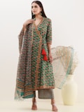 A woman stands modeling a traditional outfit featuring a long, green floral printed dress with intricate detailing and red tassels. She holds a sheer dupatta and wears matching pants. Her hair is long and straight, and she accessorizes with green earrings. A white vase with dry grass adds a decorative element to the background.