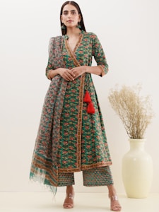 A woman is wearing a green ethnic outfit with intricate floral patterns and gold embellishments. The ensemble includes a matching kurta and palazzo pants, paired with a sheer dupatta adorned with similar designs. She is standing in a neutral-colored room beside a large, pale vase containing dried plants.