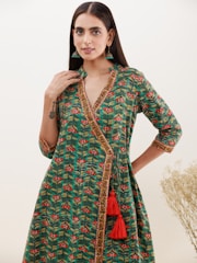 A person stands wearing a green dress with a floral pattern and intricate border detailing. The dress has a wraparound style with red tassels. The person sports long, straight hair and wears green earrings. In the background, there is a light, plain backdrop with some dried plants on the side.