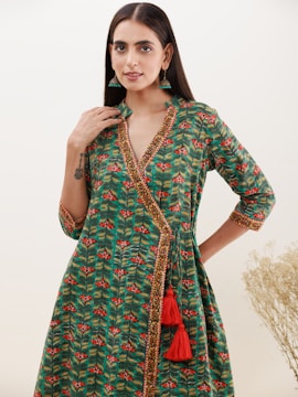 A person stands wearing a green dress with a floral pattern and intricate border detailing. The dress has a wraparound style with red tassels. The person sports long, straight hair and wears green earrings. In the background, there is a light, plain backdrop with some dried plants on the side.