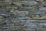 A tightly fitted stone wall made of irregularly shaped, flat pieces of slate arranged horizontally. The color palette includes various shades of gray, with subtle hints of green and brown, and the surface texture is rugged and natural.