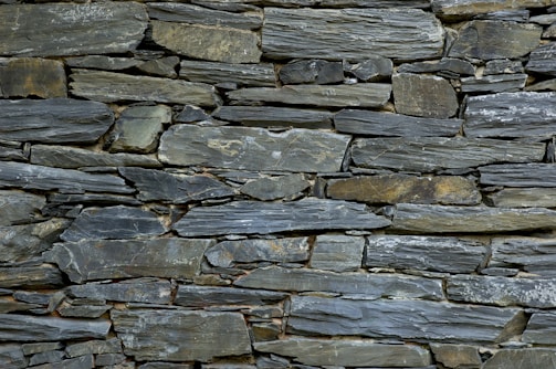 A tightly fitted stone wall made of irregularly shaped, flat pieces of slate arranged horizontally. The color palette includes various shades of gray, with subtle hints of green and brown, and the surface texture is rugged and natural.