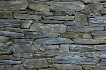 A tightly fitted stone wall made of irregularly shaped, flat pieces of slate arranged horizontally. The color palette includes various shades of gray, with subtle hints of green and brown, and the surface texture is rugged and natural.